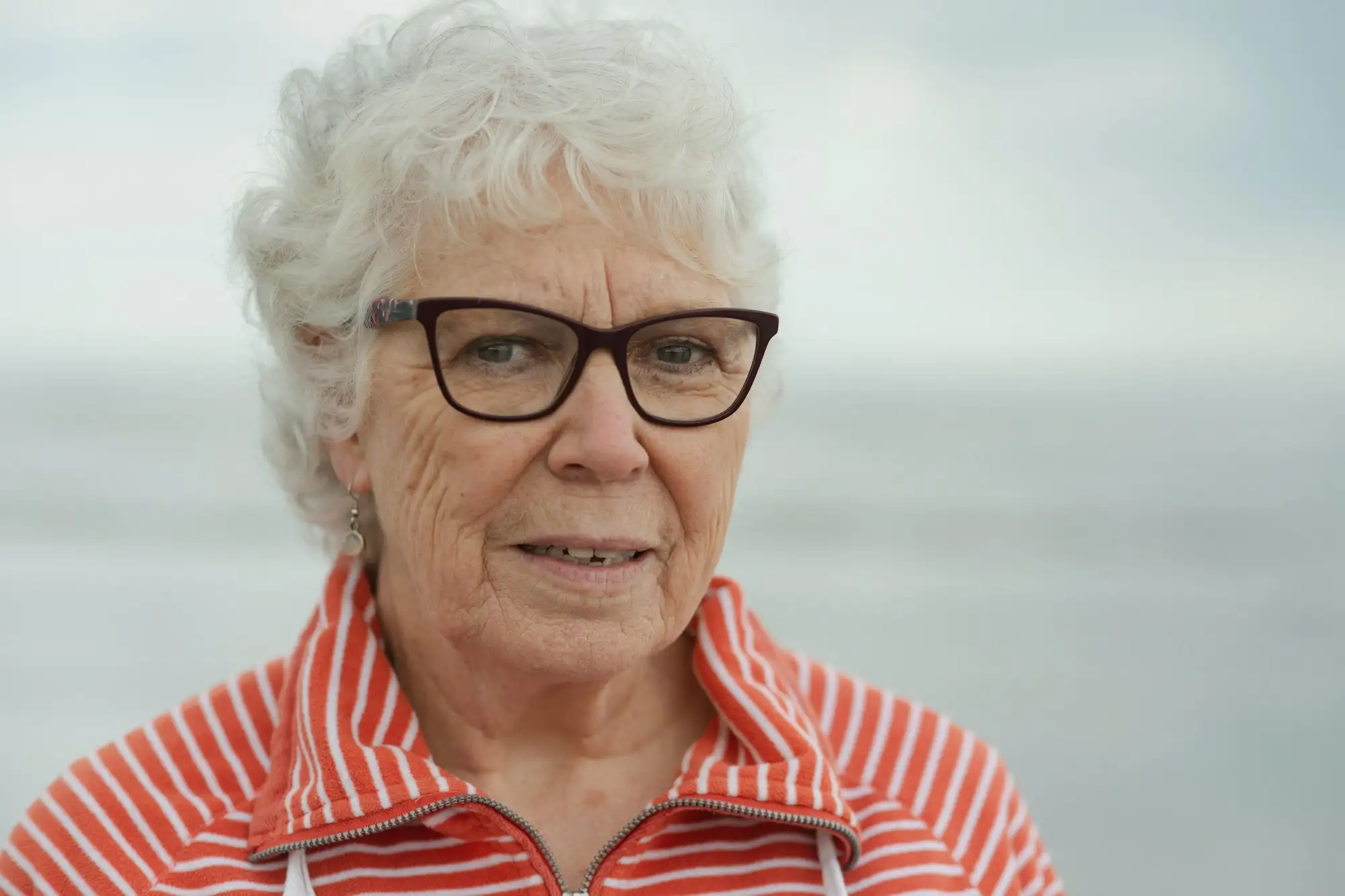 Ældre kvinde med hvidt hår og briller i rød- og hvidstribet jakke smiler ved havet