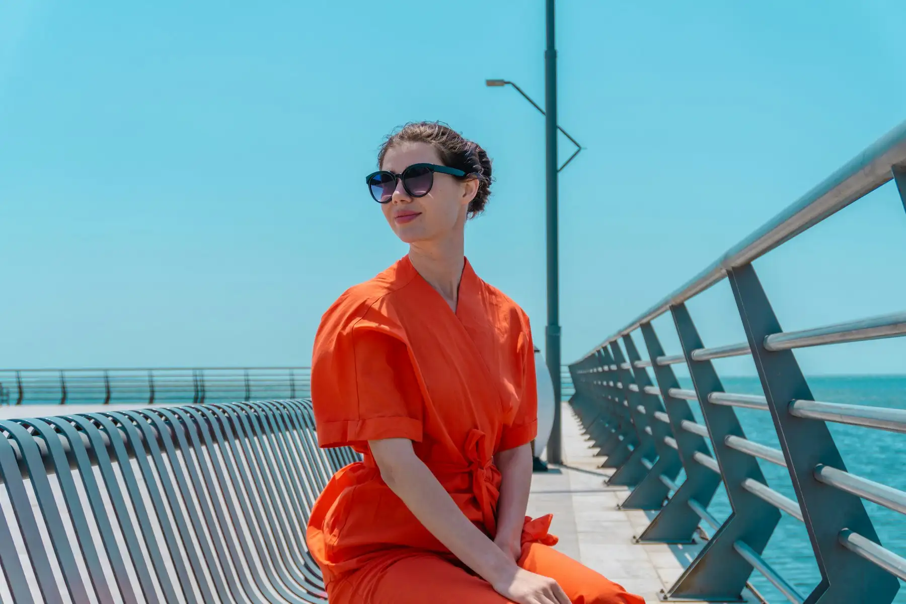 Kvinde i orange sommerkjole og solbriller sidder på en mole ved havet under en klar blå himmel