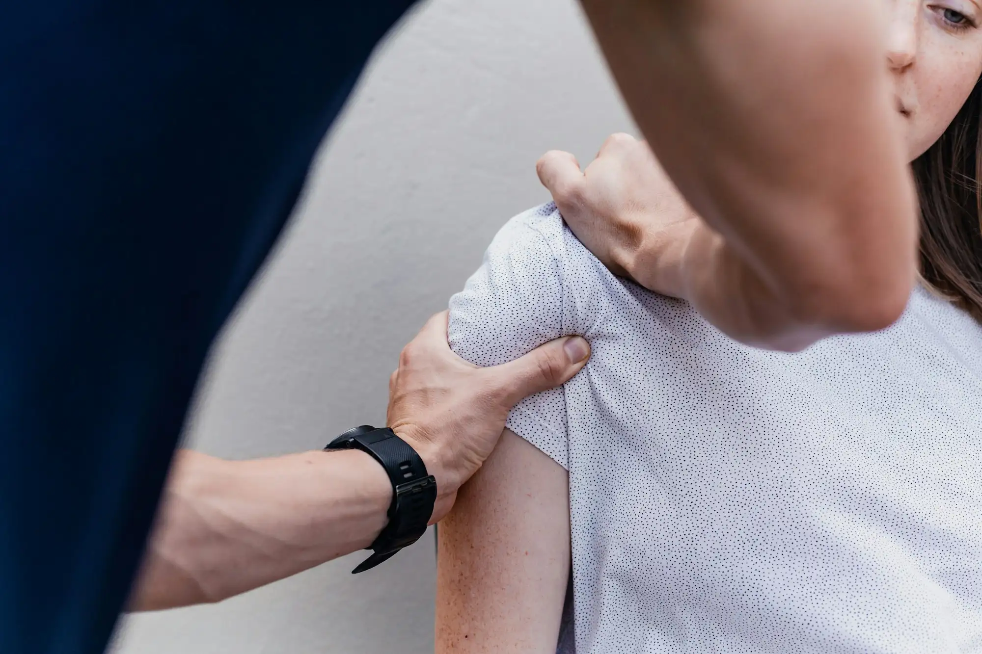 To hænder undersøger skulderen på en kvinde i hvid prikket T-shirt