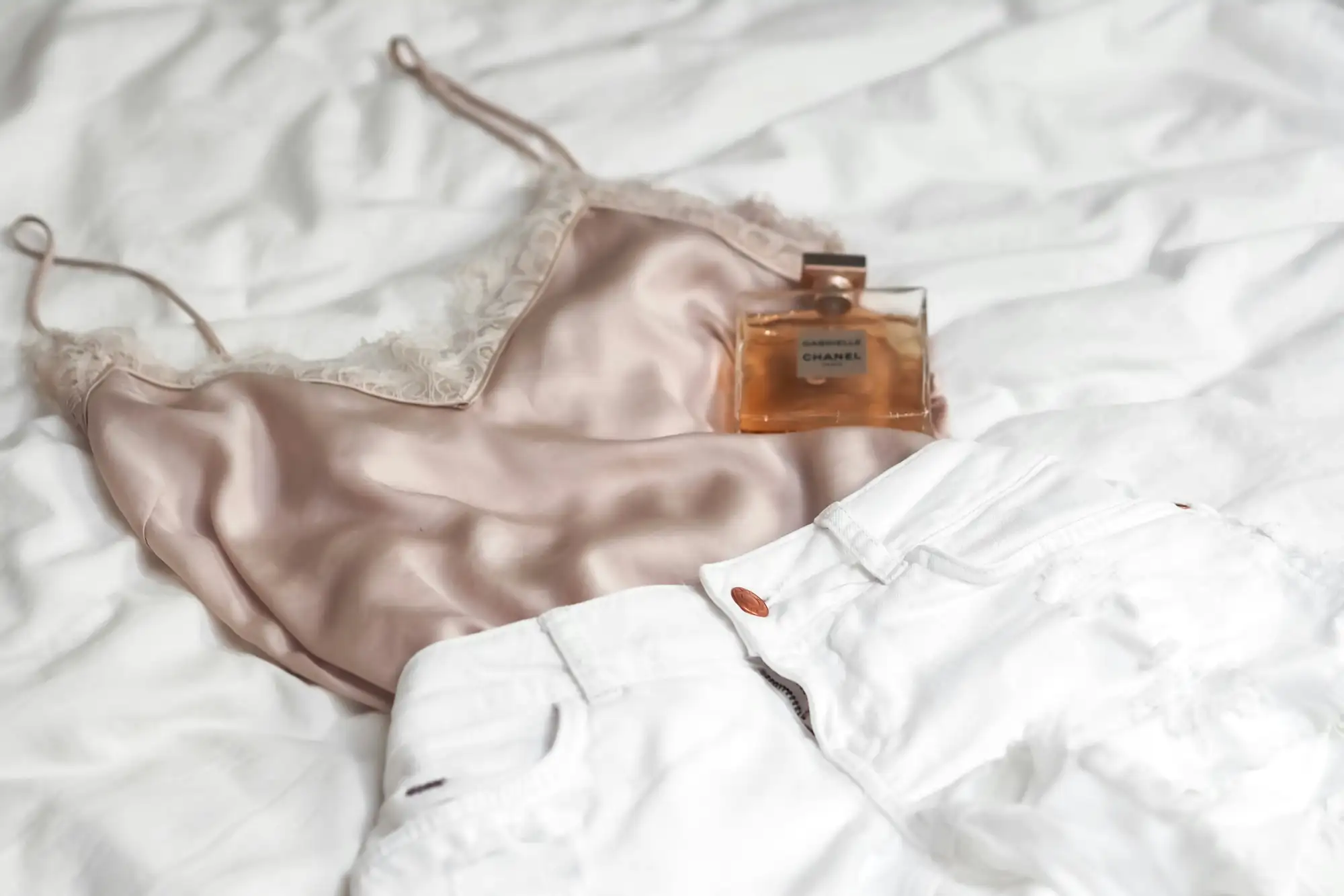 Rosa satintop med blondekant ligger på et hvidt lagen ved siden af hvide jeans og en parfumeflaske