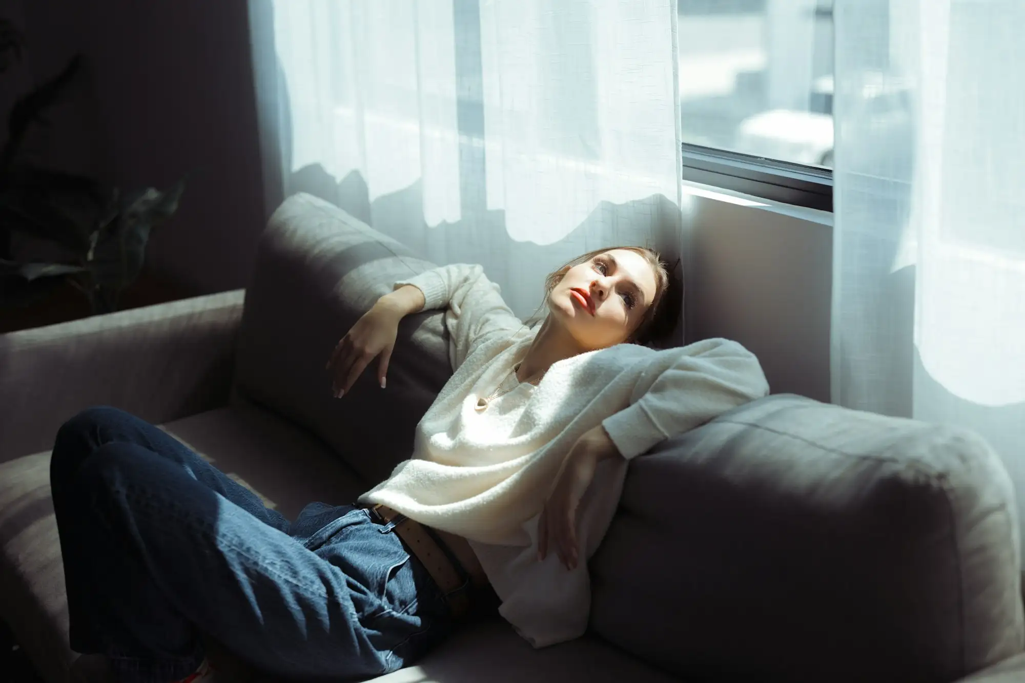 Kvinde i hvid sweater og jeans slapper af liggende på en grå sofa i sollys