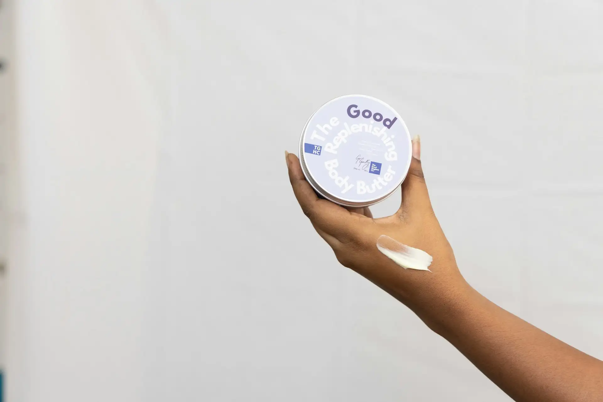 Hånd holder en dåse bodybutter med en klat creme på underarmen mod en hvid baggrund