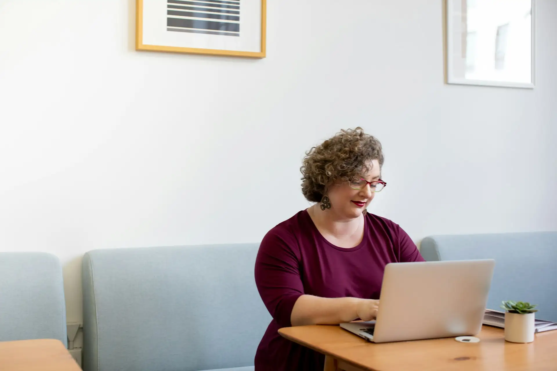 Kvinde med krøllet hår og briller sidder ret op ved et bord og arbejder på en bærbar computer