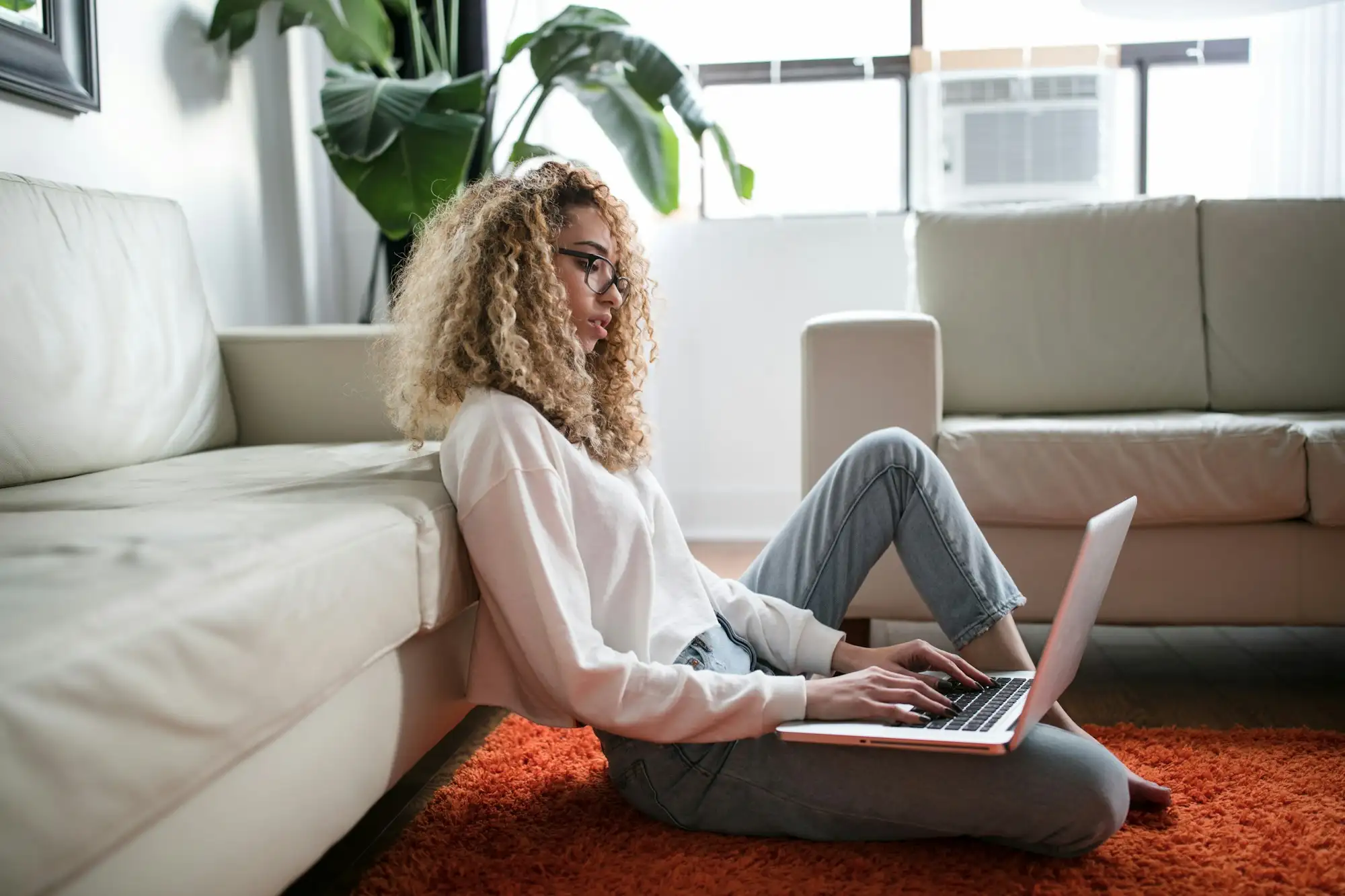 Kvinde med krøllet hår sidder på et orange tæppe op ad en sofa og skriver på en bærbar computer