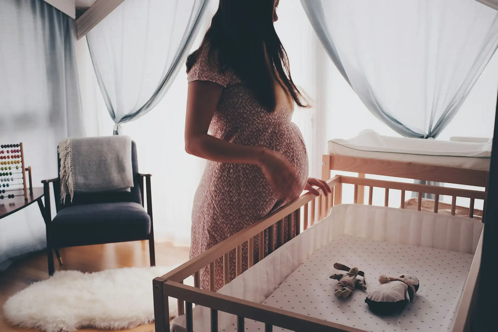 Gravid kvinde i blomstret kjole står ved en tremmeseng i et lyst børneværelse