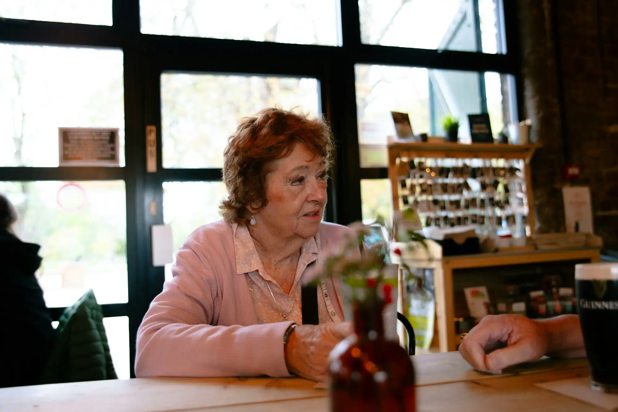 Ældre kvinde med rødt hår i lyserød cardigan sidder ved et bord på en café og taler